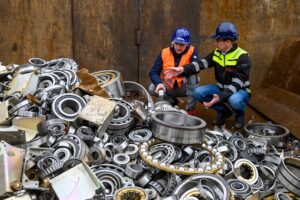Two men in safety gear squatting behind a giant pile of counterfeit ball bearings, preparing to destroy them.